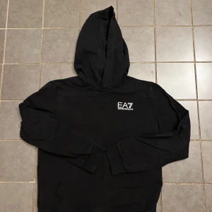 EA7 Hoodie - Helt oanvänd!! Snygg svart hoodie från Emporio Armani med EA7-logga på bröstet. Perfekt för en stilren och bekväm look. Tröjan har långa ärmar och en praktisk huva