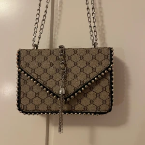 Beige axelväska med kedjedetaljer - Snygg beige axelväska med ett klassiskt mönster och silverfärgade kedjedetaljer. Väskan har en dekorativ kedja framtill och pärlor längs kanterna. Perfekt för att ge din outfit en lyxig touch.