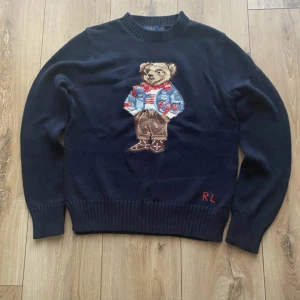 Intressekoll! Kom med bud! - Säljer en mörkblå stickad tröja från Ralph Lauren med en broderad nalle på framsidan. Tröjan har långa ärmar och ribbstickade muddar vid ärmslut och nederkant. Perfekt för en stilren och avslappnad look. Kom med bud!