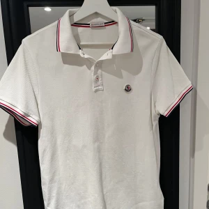 Vit pikétröja från Moncler - Säljer en stilren vit pikétröja från Moncler med klassiska ränder i rött och blått på krage och ärmslut. Tröjan har en broderad Moncler-logga på bröstet och knappar framtill. Perfekt för en avslappnad men ändå elegant look. Använd 1 gång.