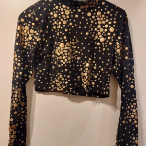 Svart och guldmönstrad croppad topp - Svart croppad topp med ett mönster av guldprickar. Toppen har långa ärmar och en hög krage, perfekt för en kväll ute. Materialet är mjukt och stretchigt för en bekväm passform. Det står XL som storlek men jag skulle säga det är en M, men det kanske beror på hur du vill att den sitter på dig.