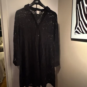 Svart spetsklänning från ICHI - Elegant svart spetsklänning från ICHI med långa ärmar och knappar framtill. Perfekt för en stilfull look med sitt vackra spetsmönster. Klänningen har en normal passform och är tillverkad i ett lätt och luftigt material.