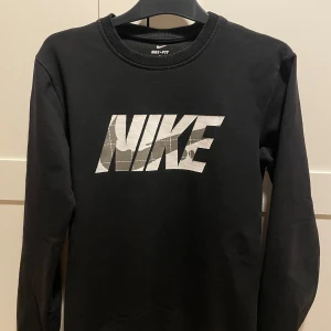 Svart långärmad tröja från Nike - Säljer en svart långärmad tröja från Nike med ett stort vitt logotryck på framsidan. Tröjan är tillverkad i Dri-FIT-material för att hålla dig torr och bekväm. Perfekt för en härlig stil och vardag. 