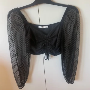 Svart topp - Snygg svart blus från NA-KD med prickigt mönster och transparenta långa ärmar. Blusen har en knytning framtill och är croppad, vilket ger en trendig look. Perfekt för att piffa upp din outfit!