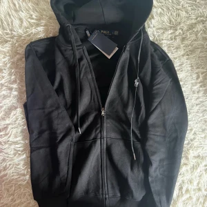 Svart hoodie från Polo Ralph Lauren - Säljer en stilren svart hoodie från Polo Ralph Lauren med dragkedja och klassisk logga på bröstet. Perfekt för en avslappnad look. Hoodien har långa ärmar och en praktisk huva med dragsko. Sälja då lillebrorsan inte tyckte om aldrig använda 