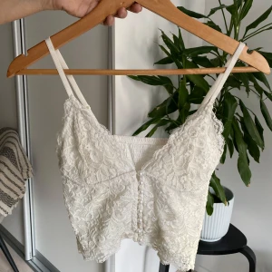 Vit spetslinne med pärldetaljer - Säljer en söt vit spetslinne med smala axelband och dekorativa pärlknappar framtill. Toppen har en vacker spetsdesign och är perfekt för varma dagar. Den är både stilren och romantisk.