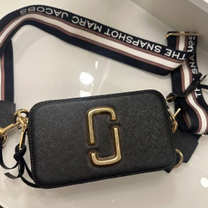 Svart axelväska från Marc Jacobs - Snygg svart axelväska från Marc Jacobs med guldiga detaljer och dubbla dragkedjor. Väskan har en bred, justerbar axelrem med text och är perfekt för att bära dina nödvändigheter. Den stilrena designen gör den till ett måste i garderoben.