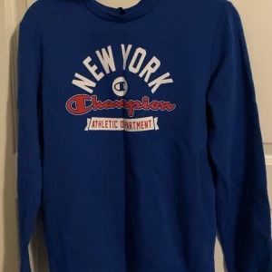 Blå långärmad tröja från Champion - Säljer en blå långärmad tröja från Champion med tryck 'New York Athletic Department' i vitt och rött på framsidan. Perfekt för en sportig look eller avslappnad stil. Tröjan har en rund halsringning och är gjord i ett mjukt material.