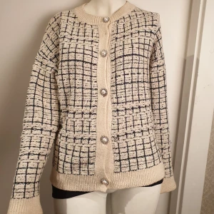 Crémevit och mörkblå rutig kofta med pärlknappar - Snygg vit kofta med mörkblå rutmönster och dekorativa pärlknappar. Perfekt för att ge en elegant touch till din outfit. Koftan har långa ärmar och ribbade kanter.