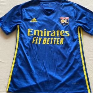 Lyon fotbollströja+byxor från Adidas - Snygg Lyon fotbollströja från Adidas med gula detaljer och korta ärmar. Tröjan har ett tryck av Emirates Fly Better och klubbmärke på bröstet. Tillverkad i Climalite-material för optimal komfort.