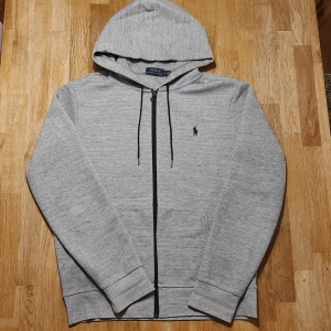 Grå hoodie från Ralph Lauren - Säljer denna riktigt snygga hoodie från Ralph Lauren. Gråmelerad med svart logga och svarta detaljer. Glatt lyxig känsla på tyget. Superskick, inga defekter! Stl M, upplevs true to size, se mått. Armhåla till armhåla 57cm, bredd längst ner 46cm, längd från mitt på axeln 65cm.
