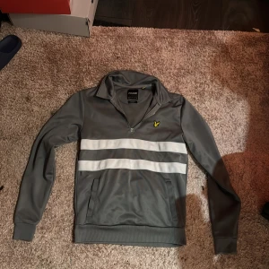 Grå tröja med dragkedja från Lyle & Scott - Säljer en grå tröja från Lyle & Scott med dragkedja och två vita ränder över bröstet. Tröjan har en avslappnad passform och långa ärmar. Perfekt för en stilren och bekväm look.
