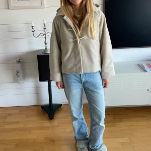 Beige kappa med huva - Säljer en stilren beige kappa med huva och knäppning framtill. Jackan har en avslappnad passform och är perfekt för kyligare dagar. Den är tillverkad i ett mjukt material som ger en bekväm känsla. Passar bra till jeans och sneakers för en casual look.
