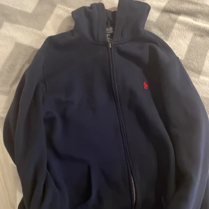 Mörkblå hoodie från Polo Ralph Lauren - Säljer en snygg mörkblå hoodie från Polo Ralph Lauren med dragkedja och röd broderad logga på bröstet. Perfekt för en avslappnad stil. Den har en bekväm passform och huva för extra mysfaktor. Storlek xs . Xl i barnstolek. Nyskick använt endast 1 gång 