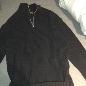 Svart stickad tröja från Smog - Säljer en svart stickad tröja från Smog med dragkedja vid halsen. Tröjan har långa ärmar och en ribbad struktur som ger en stilren look. Perfekt för kyligare dagar.