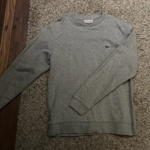 Lacoste Sweatshirt - Säljer en stilren grå sweatshirt från Lacoste med det klassiska krokodilemblem på bröstet. Tröjan har långa ärmar och rund halsringning, perfekt för en avslappnad look. 100% Bomull