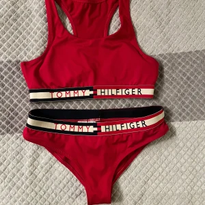 Röd bikini från Tommy Hilfiger - Snygg röd bikini från Tommy Hilfiger med sportig design. Stlk: XS. Överdelen har en bred resår med logotyp och brottarrygg, medan underdelen har en matchande resår med logotyp. Perfekt för en dag på stranden eller vid poolen.