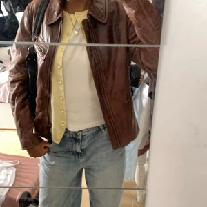 Brun äkta skinnjacka från UNIQLO - Säljer en snygg brun skinnjacka med röd quiltad foder. Jackan har en dragkedja framtill och flera praktiska fickor med dragkedjor. Perfekt för en cool och stilren look. Äkta läder