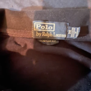 Svart pikétröja från Ralph Lauren - Säljer en klassisk svart pikétröja från Ralph Lauren med långärmad design. Tröjan har en röd broderad logotyp på bröstet och knappar vid kragen. Perfekt för en stilren look.