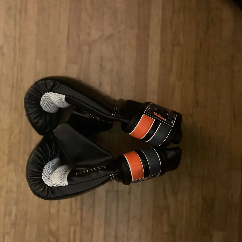 Snygga svarta boxningshandskar från Fighter med orange och grå detaljer. Perfekta för träning, de har en robust design och breda kardborreband för extra stöd. Handskarna har en ventilerad insida för bättre komfort under intensiva pass.. Asusteet.
