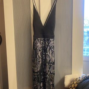 Svart glittrig långklänning - Elegant svart långklänning med djup v-ringning och smala axelband. Kjolen är täckt av ett glittrigt mönster i silver och guld som ger en lyxig känsla. Perfekt för en glamorös kväll.
