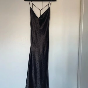 Svart långklänning med öppen rygg - Elegant svart långklänning med tunna axelband och en stilren öppen ryggdesign. Klänningen har en slät yta som ger ett lyxigt intryck. Perfekt för en kväll ute eller en speciell tillställning. Har toppror men ändå väldigt fin!