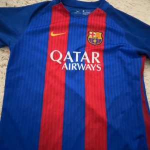 Barcelona tröja - Säljer en klassisk FC Barcelona fotbollströja från Nike i blått och rött med klubbens emblem på bröstet. Tröjan har korta ärmar och ett randigt mönster. Perfekt för fans av laget!