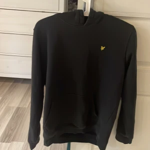 Svart hoodie från Lyle & Scott Junior - Säljer en stilren svart hoodie från Lyle & Scott . Den har en klassisk design med en liten gul logga på bröstet och en bekväm huva. Perfekt för en avslappnad look.