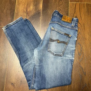 Blå jeans från Nudie - Säljer ett par snygga blå jeans från Nudie i bra skick. Storlek W32, L30, De har en klassisk femficksdesign med en cool broderad detalj på bakfickan. Jeansen är i en straight passform och har en dragkedja. Perfekta för både vardag och fest!
