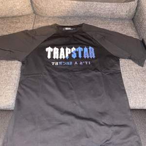 Säljer en svart t-shirt från Trapstar med tryck i vitt och blått på framsidan. T-shirten har korta ärmar och en rund halsringning. Perfekt för en casual look eller streetwear-stil. Passar bra till jeans eller shorts!