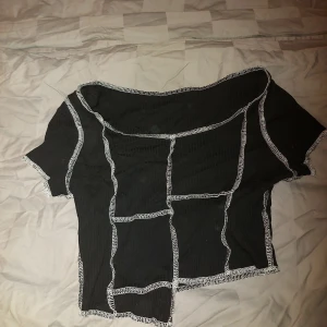Svart topp från SHEIN - Säljer en svart topp från SHEIN i storlek S. Den har en unik design med vita sömmar som skapar ett rutmönster. Toppen är kortärmad och har en offshoulder-stil som ger en trendig look. Perfekt för både vardag och fest!