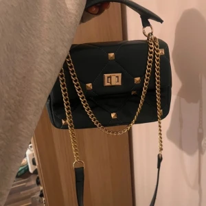 Väska  - Fin väska i riktigt bra skick! Väskan är grönblå med guldiga detaljer, en justerbar kedja och ett handtag som går att ta av. Man kan alltså bära väskan på olika sätt! Priset kan variera beroende på hur många som är intresserade.