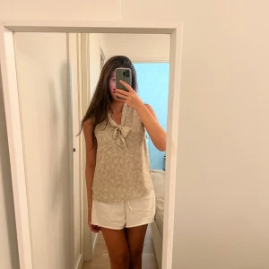 Beige blommig ärmlös topp - Säljer en söt beige ärmlös topp med blommigt mönster. Den har en knytning framtill som ger en extra touch. Perfekt för sommardagar eller en avslappnad vårlook. Toppen är i mycket bra skick och passar till både shorts och kjol.