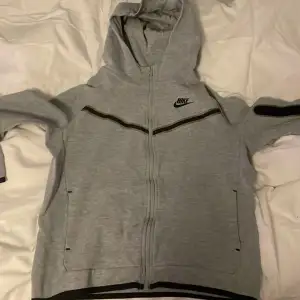 Säljer en grå hoodie från Nike i bra skick. Den har en dragkedja framtill och en klassisk Nike-logga på bröstet. Perfekt för höst och vår, med långa ärmar och en bekväm huva. Passar både till träning och vardagsbruk.