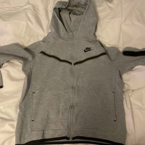 Grå Nike tech  - Säljer en grå hoodie från Nike i bra skick. Den har en dragkedja framtill och en klassisk Nike-logga på bröstet. Perfekt för höst och vår, med långa ärmar och en bekväm huva. Passar både till träning och vardagsbruk.