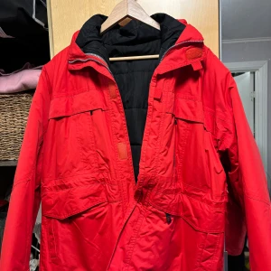 Röd vinterjacka från Silanta - Säljer en snygg och praktisk röd vinterjacka från Silanta i storlek XL. Jackan har flera fickor och en dragkedja framtill. Perfekt för kalla vinterdagar med sin varma vaddering. Passar både till vardags och utomhusaktiviteter. Samt att det är uttagbart foder i den så att den går att använda som bara vindjacka