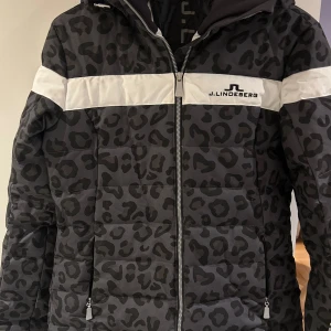Svart leopardmönstrad jacka från J.Lindeberg - Säljer en svart/grå leopardmönstrad jacka från J.Lindeberg. Jackan har en dragkedja framtill och en vit rand över bröstet och ryggen. Perfekt för att hålla sig varm och snygg under kyliga dagar. Passar både till vardags och i skidbacken