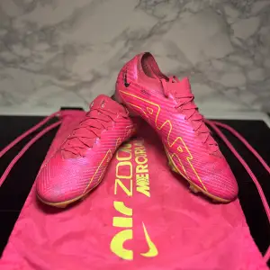 Säljer ett par grymma Nike Air Zoom Mercurial fotbollsskor i en cool rosa färg med gula detaljer. Skorna har snörning och är i syntetmaterial, perfekta för fotbollsspel på gräs. Sulan har bra grepp och är designad för snabbhet och kontroll. De är i mycket bra skick och redo för nästa match!. (Priset kan diskuteras)