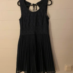 Mörkblå spetsklänning från Vero Moda - Säljer en superfin mörkblå klänning från Vero Moda. Den har en vacker spetsdetalj upptill och ett plisserat kjolparti som ger en elegant look. Klänningen är ärmlös och har en söt knytning i ryggen. Perfekt för festliga tillfällen!