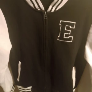 Svart och vit bomberjacka med bokstaven E - Säljer en snygg svart och vit bomberjacka med en stor bokstav E på bröstet. Jackan har dragkedja framtill och långa ärmar. Perfekt för en cool och avslappnad stil!