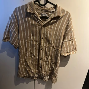 Beige och vit randig skjorta från Jack & Jones - Säljer en beige och vit randig skjorta från Jack & Jones. Skjortan har korta ärmar och knappar framtill. Perfekt för en avslappnad stil.