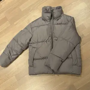 Säljer en stilren grå pufferjacka från BikBok i återvunnen polyester. Jackan har en dragkedja framtill och är perfekt för kyligare dagar. Den har en hög krage och långa ärmar för extra värme.
