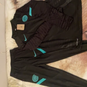 Svart träningsset från Nike med Inter-logga - Säljer ett svart träningsset från Nike med Inter-logga i turkos. Setet består av en långärmad tröja och matchande byxor. Perfekt för fotbollsträning eller annan sportaktivitet. Nike-loggan är också i turkos, vilket ger en snygg kontrast mot det svarta tyget.