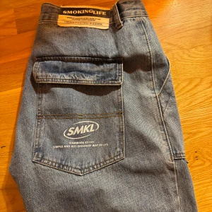 Blåa jeans Thrifted  - Säljer ett par stora blå jeansbyxor från SMKL med en avslappnad passform. Byxorna har en praktisk ficka med lock på sidan och en tydlig logga på baksidan. Perfekta för en casual look! 