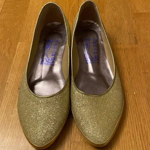 Glittriga guldiga ballerinaskor - Snygga glittriga ballerinaskor i guld med låg klack. Perfekta för att lägga till lite glamour till din outfit.