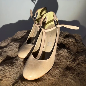 Beige pumps med rem - Eleganta beige pumps med rund tå och smal klack. Skorna har en justerbar rem runt ankeln för extra komfort och stil.