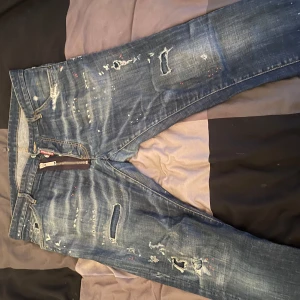 Blå jeans från Dsquared2 - Snygga blå jeans från Dsquared2 med slitna detaljer och färgstänk. De har en dragkedja och knappar framtill samt en läderpatch med logotyp på baksidan. Perfekta för en trendig look. Öppen för att gå ner i pris via snabbaffär🌟