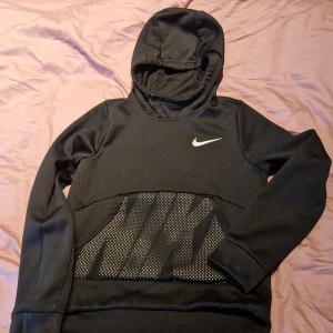 Svart hoodie från Nike - Säljer en svart hoodie från Nike med Dri-FIT-teknologi. Den har en vit Nike-logga på bröstet och ett mönstrat parti framtill. Perfekt för sportiga aktiviteter eller en avslappnad stil.