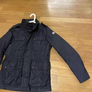  Moncler field jacka - Snygg mörkblå jacka från Moncler med dragkedja och knappar. Jackan har flera praktiska fickor och en hög krage. Perfekt för kyligare dagar. - modell namn Christian - size 4- mycket fint skick inga flaws förutom att klädhängaren är lite lös -