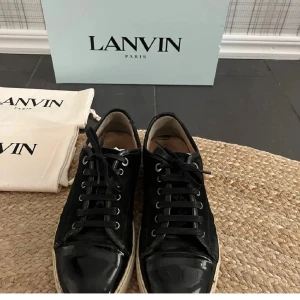Svarta sneakers från Lanvin - Snygga svarta sneakers från Lanvin med snörning och en stilren design. Skorna har en blank yta och en vit sula som ger en klassisk look. Perfekta för en stilmedveten garderob. Defekt på bild 3,4,5 box ingår om man frågar dust bag ingår ej
